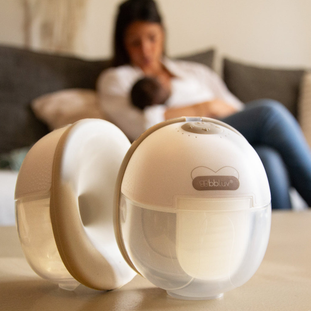 bblüv electric breast pump on a table with a mother breastfeeding her baby in the background
